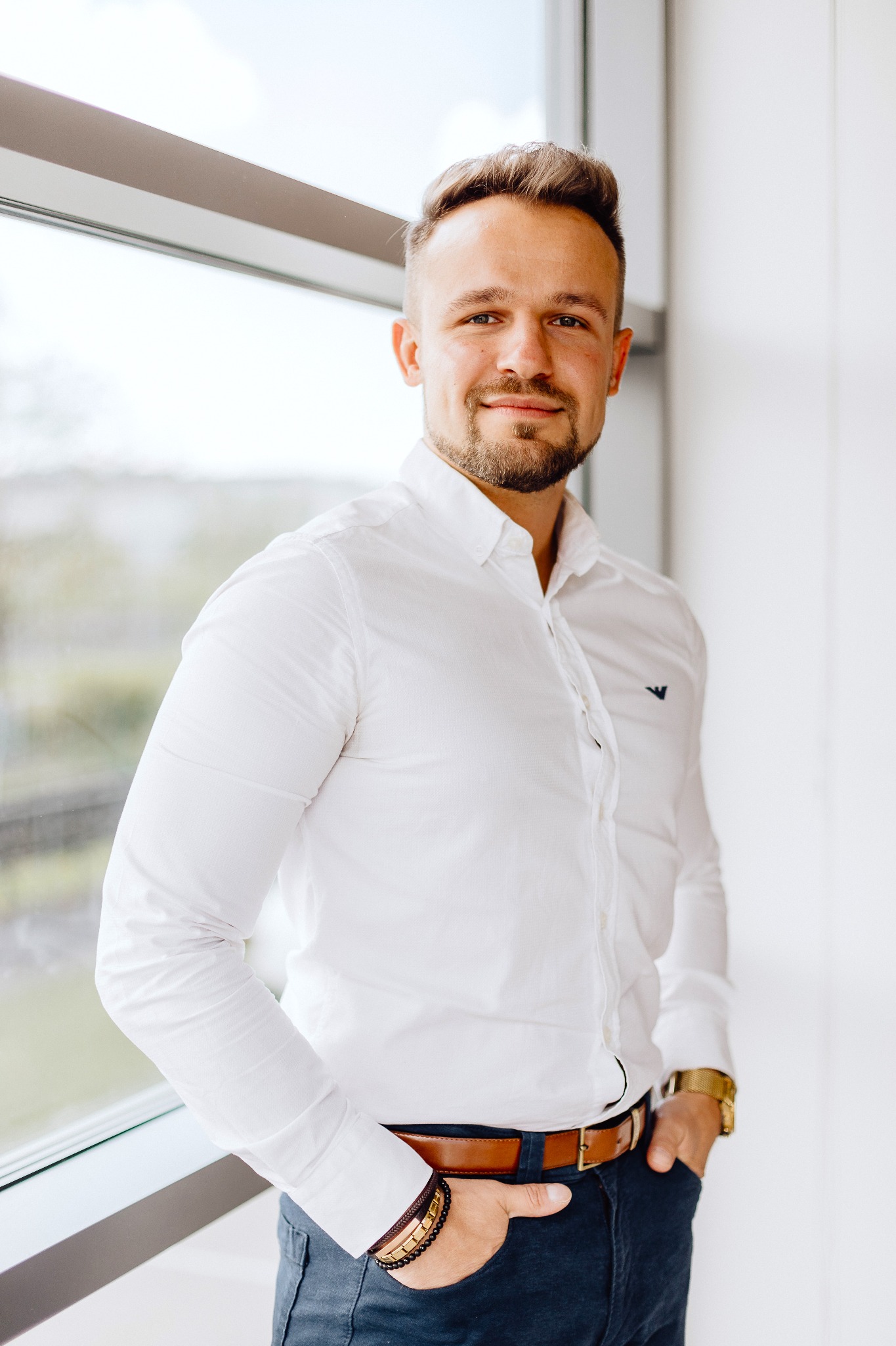 Portret uśmiechniętego mężczyzny w białej koszuli, stojącego przy oknie z ręką w kieszeni spodni.