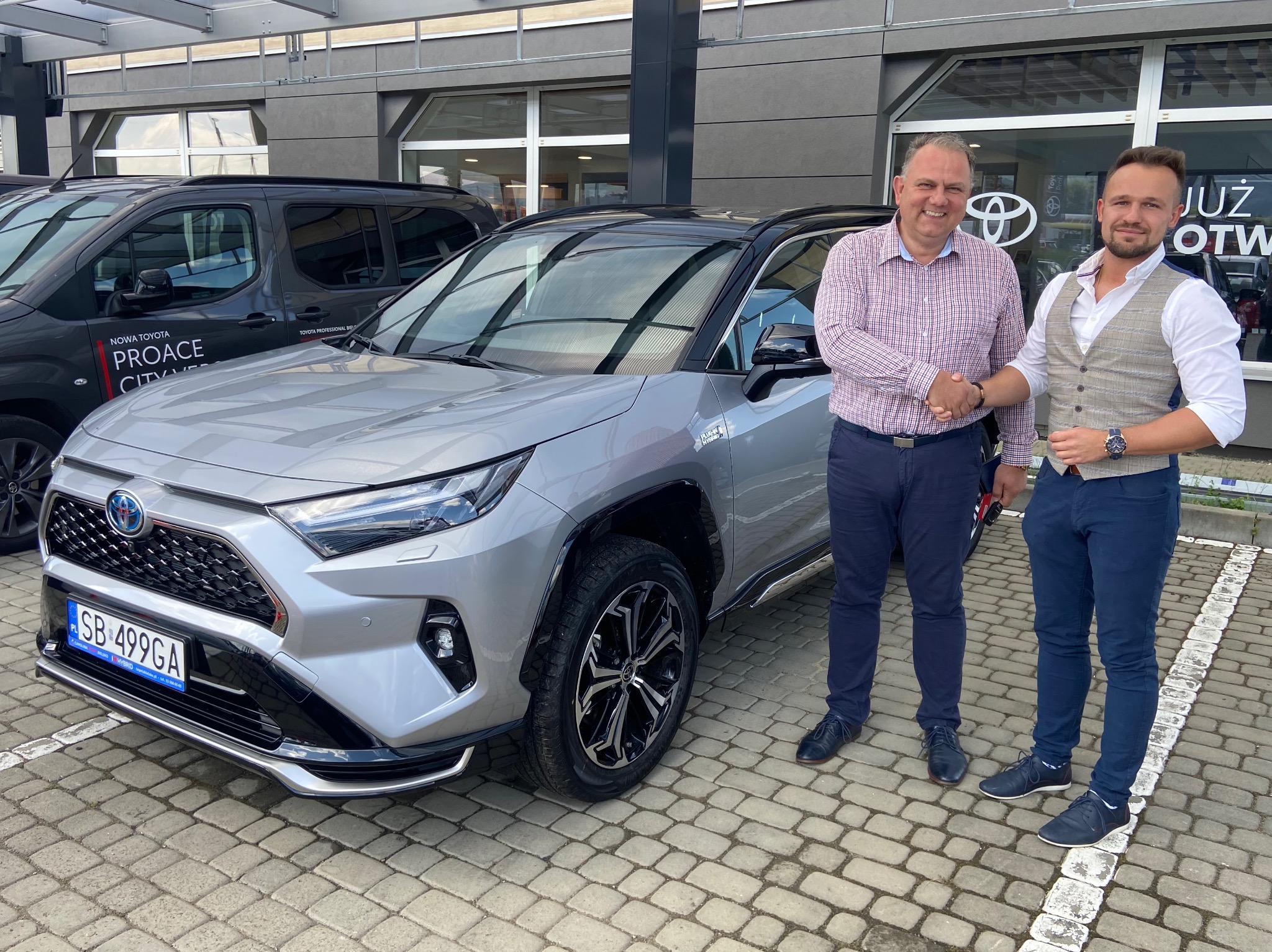 Srebrna Toyota RAV4 na tle salonu samochodowego, dwaj mężczyźni w garniturach uściskują sobie dłonie przed pojazdem, widoczna tablica rejestracyjna.