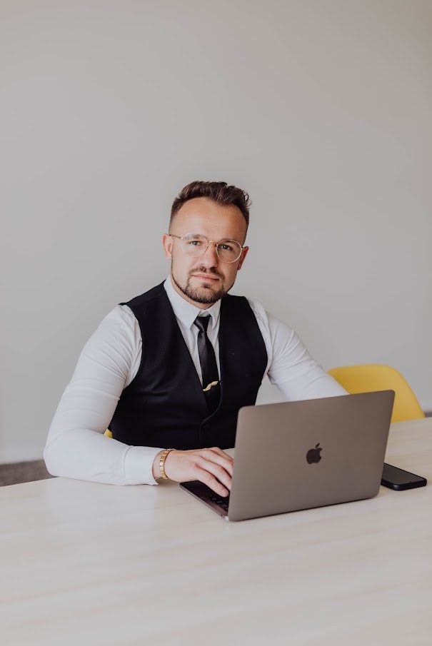 Mężczyzna w okularach, kamizelce i z krawatem siedzi przy biurku i pracuje na laptopie, obok leży smartfon.