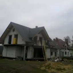 Budowa domu jednorodzinnego w trakcie krycia dachu, widoczna częściowo ułożona dachówka w kolorze grafitowym, surowy stan elewacji, drabina oparta o ścianę, pochmurne niebo w tle.