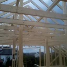 Surowa drewniana konstrukcja więźby dachowej w trakcie montażu, widok z wnętrza budynku na tle błękitnego nieba i zabudowy jednorodzinnej.