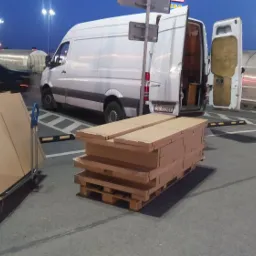 Biały van z otwartymi tylnymi drzwiami na parkingu, obok wózka z kartonami i palety z kartonami, sugerujące załadunek lub rozładunek towaru w godzinach wieczornych.