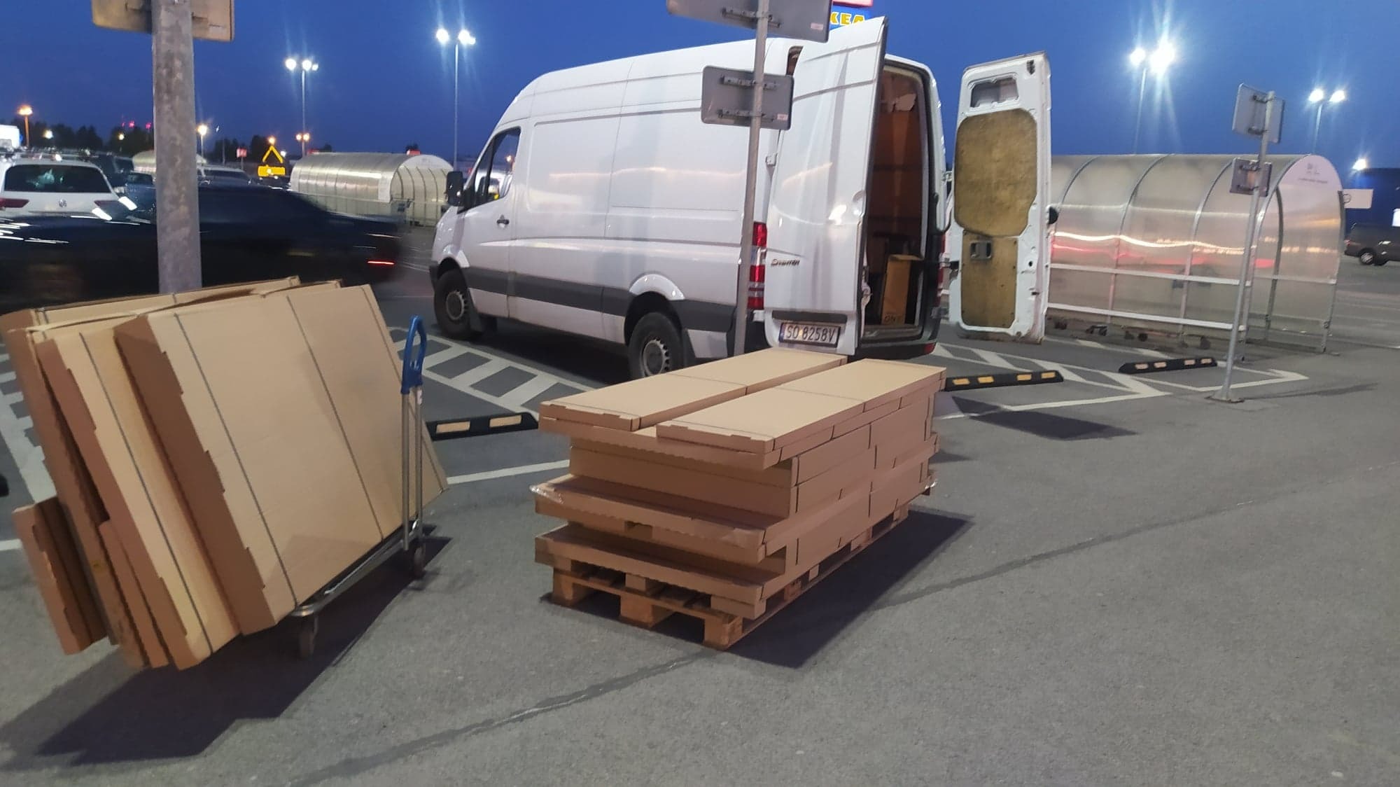 Biały van z otwartymi tylnymi drzwiami na parkingu, obok wózka z kartonami i palety z kartonami, sugerujące załadunek lub rozładunek towaru w godzinach wieczornych.