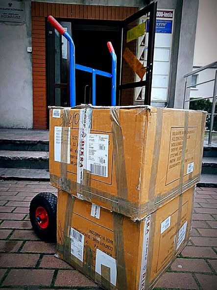 Dwa kartony zabezpieczone taśmą na niebieskim wózku transportowym z czerwonymi uchwytami przed wejściem do budynku z szyldem 'Kancelaria Notarialna'.