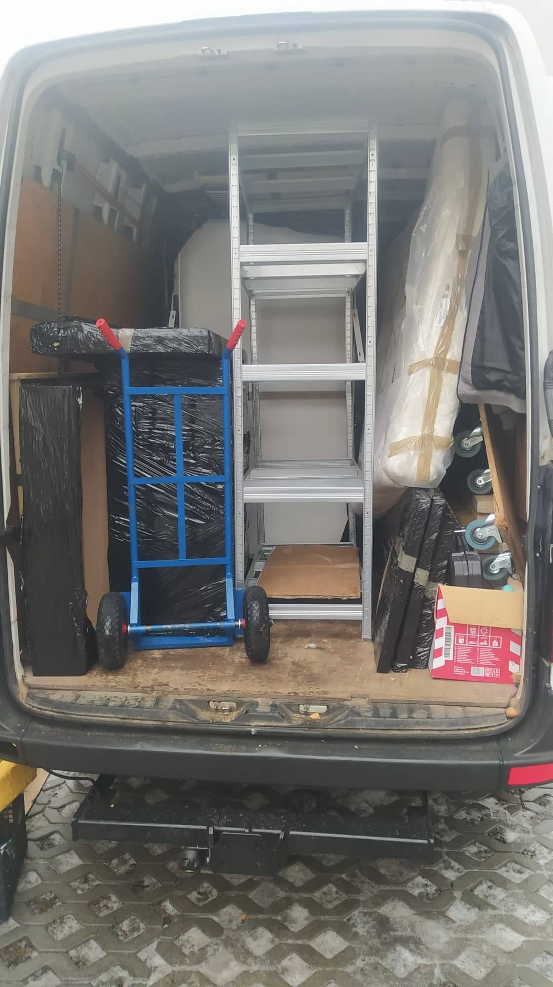 Wnętrze busa załadowane meblami i sprzętem, zabezpieczonym folią, z niebieskim wózkiem transportowym i metalowym regałem, przygotowane do transportu.