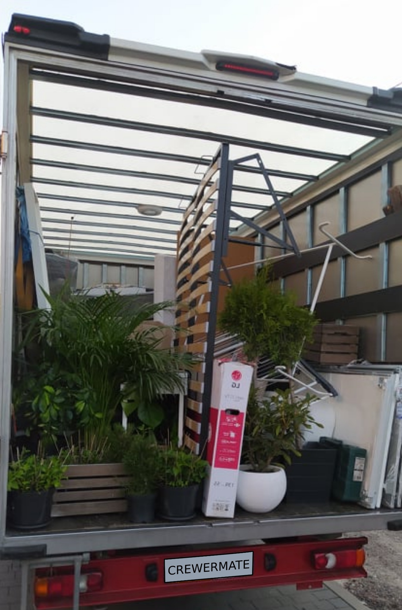 Wnętrze furgonetki załadowane roślinami doniczkowymi, elementami łóżka, kartonami i innymi przedmiotami podczas transportu, widok od tyłu pojazdu.