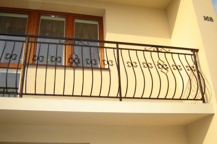 Czarna, metalowa balustrada balkonowa z dekoracyjnymi elementami w kształcie stylizowanych ślimaków i rombów, zamontowana na jasnym balkonie budynku z beżową elewacją i drewnianą ramą okienną.