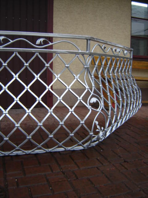 Kuta, metalowa balustrada balkonowa z motywem rombów i dekoracyjnymi elementami roślinnymi, widoczna z niskiej perspektywy na tle ceglanej posadzki i elewacji budynku.