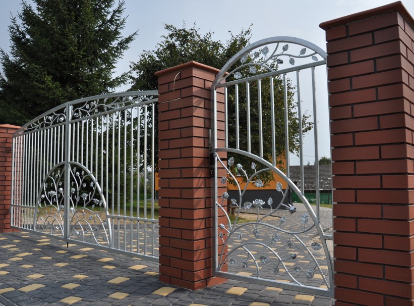 Metalowa brama wjazdowa z dekoracyjnymi elementami roślinnymi, osadzona na ceglanych słupkach, prowadząca na posesję z widocznym domem i drzewami w tle.