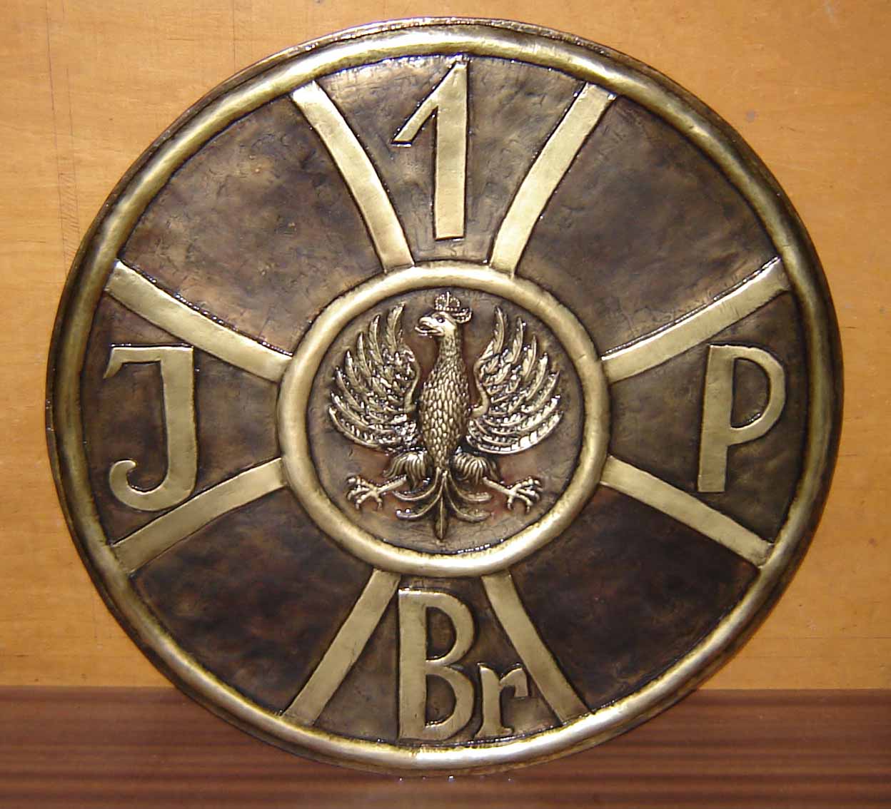 Metalowa płaskorzeźba w kształcie koła z orłem w koronie, literami J, P, Br i cyfrą 1, stylizowana na kowalstwo artystyczne.
