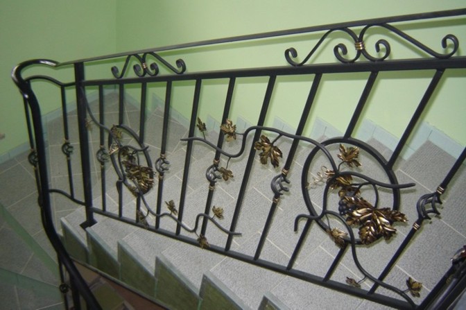 Kuta balustrada schodowa z motywem winorośli, czarne metalowe elementy z dekoracyjnymi, złotymi detalami liści, widok z góry na schody wyłożone szarymi płytkami.