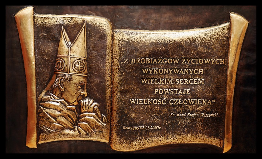 Metalowa płaskorzeźba przedstawiająca postać Kardynała Stefana Wyszyńskiego i cytat, stylizowana na otwartą księgę w odcieniach złota i brązu.