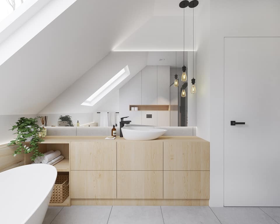 Jasna łazienka na poddaszu ze skośnym sufitem, umywalką nablatową, drewnianą szafką pod umywalką, wanną, lustrem na całą ścianę i wiszącymi lampami z żarówkami Edisona.