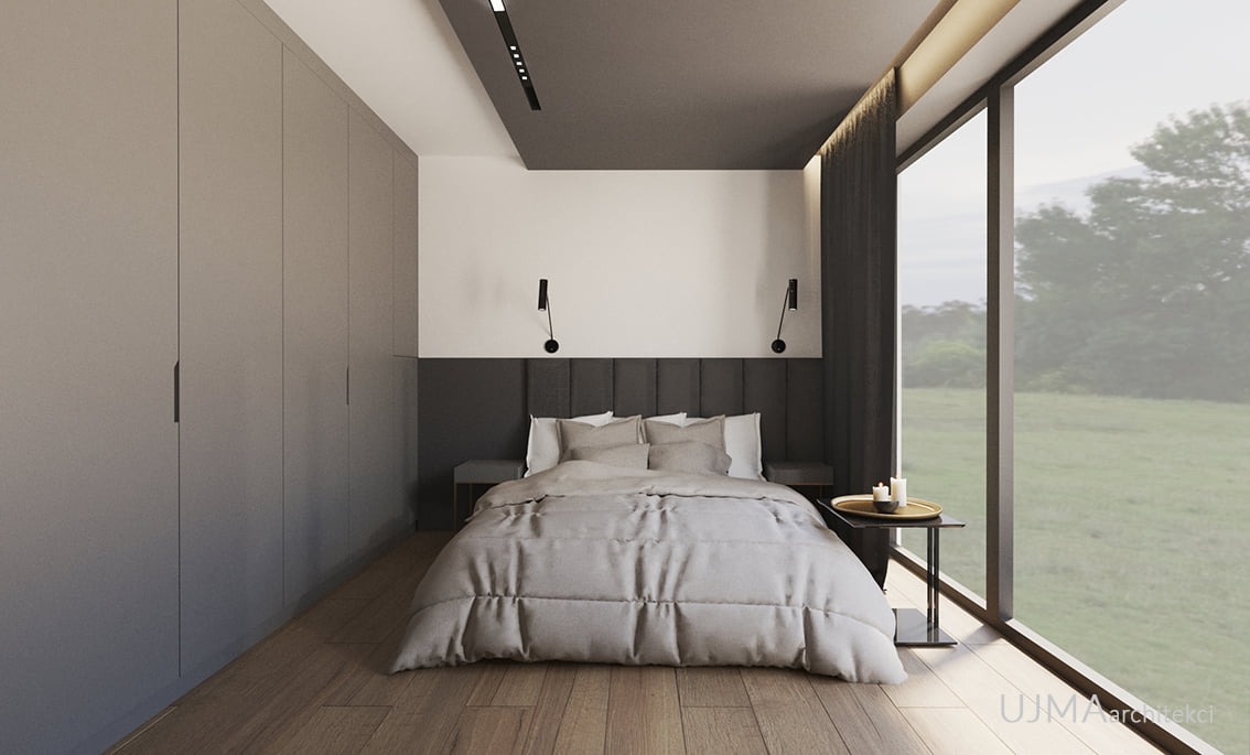 Nowoczesna sypialnia z dużym oknem wychodzącym na zielony krajobraz, szara tapicerowana ściana za łóżkiem, minimalistyczne lampki ścienne i szafa w zabudowie po lewej stronie.