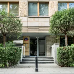 Wejście do budynku z dwoma kulistymi drzewami, niskim żywopłotem i czarnymi słupkami ograniczającymi przejście.