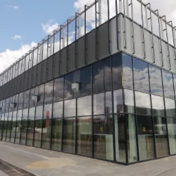 Nowoczesny budynek biurowy z szklaną fasadą odbijającą chmury i ażurową konstrukcją na dachu, widoczny z perspektywy chodnika.