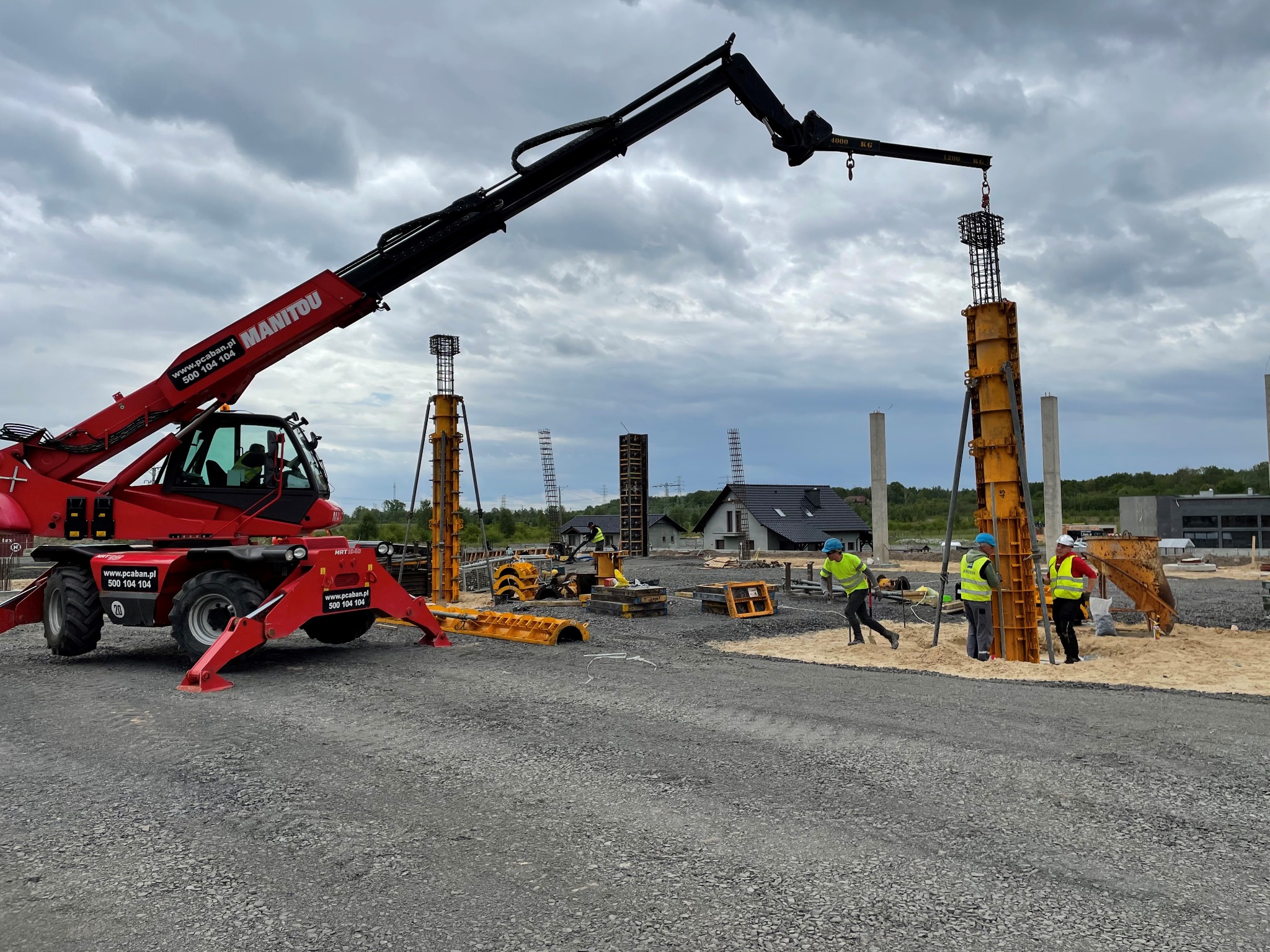 Montaż zbrojenia słupa fundamentowego przy użyciu dźwigu teleskopowego Manitou, pracownicy w kaskach i kamizelkach odblaskowych nadzorują proces na placu budowy.