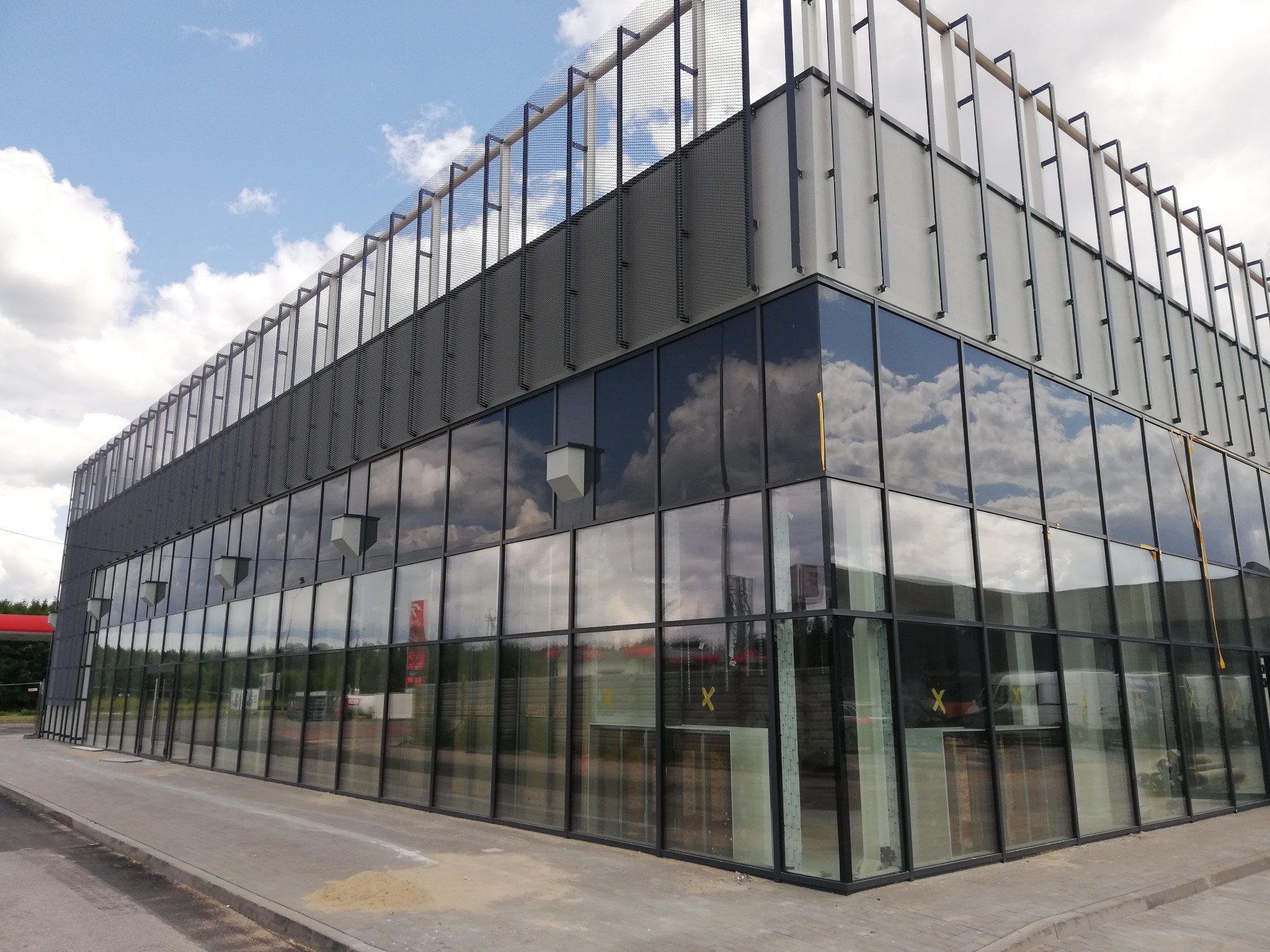 Nowoczesny budynek biurowy z szklaną fasadą odbijającą chmury i ażurową konstrukcją na dachu, widoczny z perspektywy chodnika.