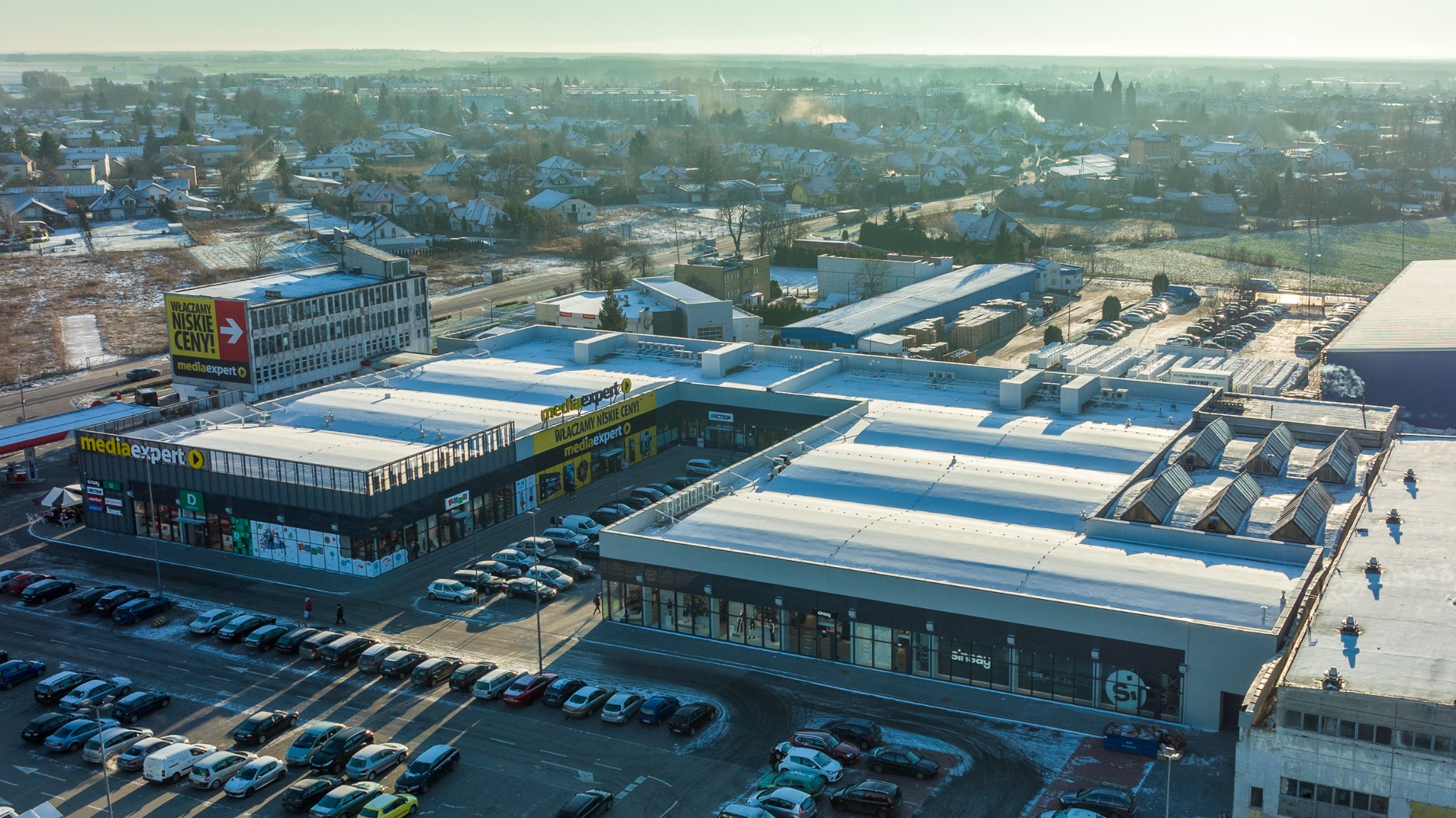Widok z góry na kompleks handlowy z parkingiem pokrytym cienką warstwą śniegu, w tle zabudowa miejska i lekka mgła