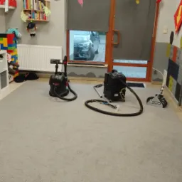 Profesjonalny sprzęt do czyszczenia dywanów ustawiony na szarym dywanie w pokoju dziecięcym z zabawkami i kolorowymi dekoracjami na ścianach.