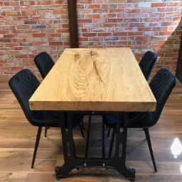 Stół z blatem z jasnego drewna o wyraźnym usłojeniu i czarną metalową podstawą w stylu industrialnym, otoczony czterema czarnymi krzesłami na tle ściany z czerwonej cegły.