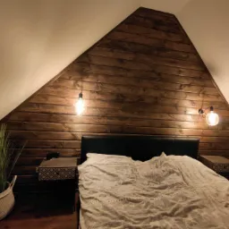 Pokój na poddaszu z drewnianą ścianą szczytową, oświetlony żarówkami w stylu retro, łóżko z czarnym zagłówkiem i wzorzystą pościelą, stoliki nocne z geometrycznym wzorem i dekoracyjna trawa...