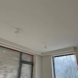 Wnętrze w trakcie remontu: świeżo pomalowany sufit, zabezpieczone okna folią malarską, wystające kable oświetleniowe.