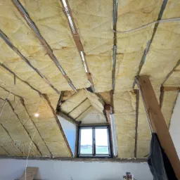 Poddasze w trakcie ocieplania, widoczne wełna mineralna między krokwiami, metalowe profile, sznurki mocujące i okno dachowe z widokiem na krajobraz.