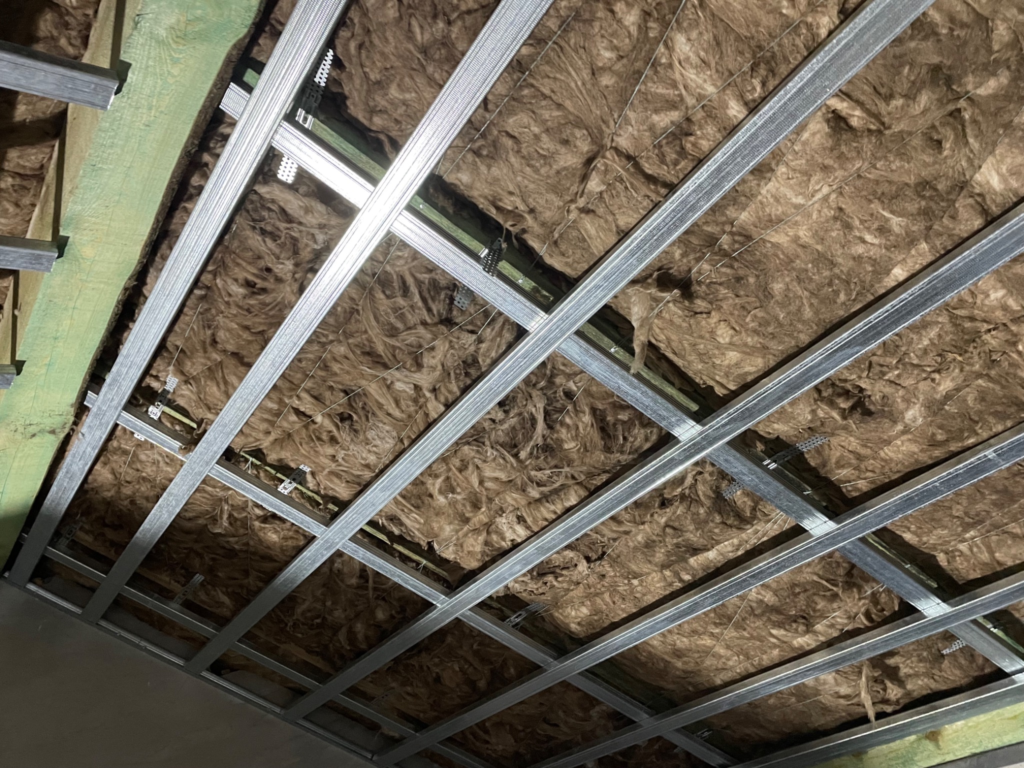 Sufit podwieszany z wełną mineralną między profilami, widoczne elementy mocujące i konstrukcja nośna z metalowych profili.