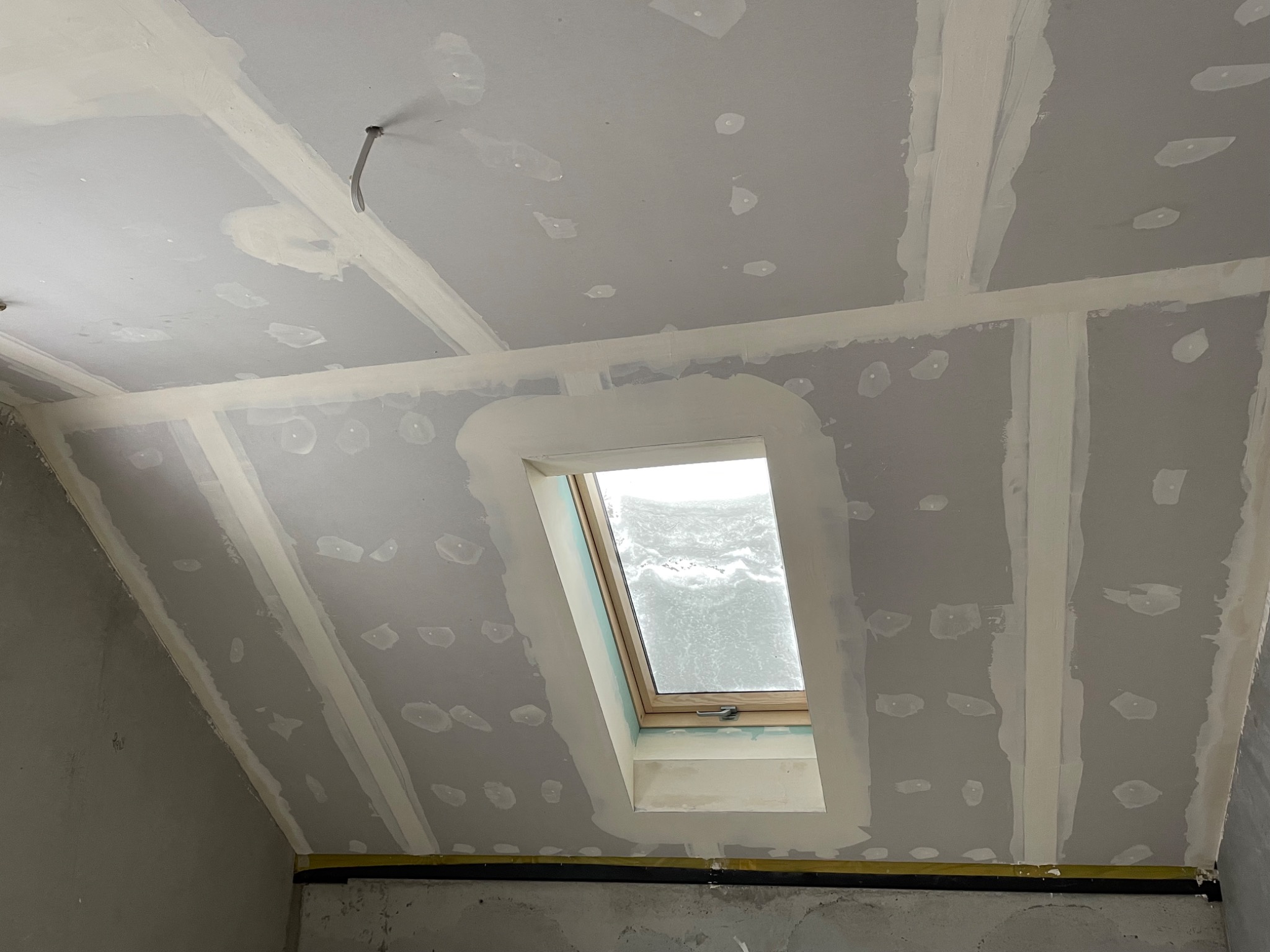 Poddasze w trakcie wykańczania: zamontowane okno dachowe, widoczne płyty gipsowo-kartonowe z zaszpachlowanymi łączeniami oraz wystający kabel.