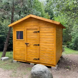 Drewniana altana ogrodowa z małym oknem i czarnymi zawiasami, ustawiona na betonowych bloczkach w otoczeniu drzew.