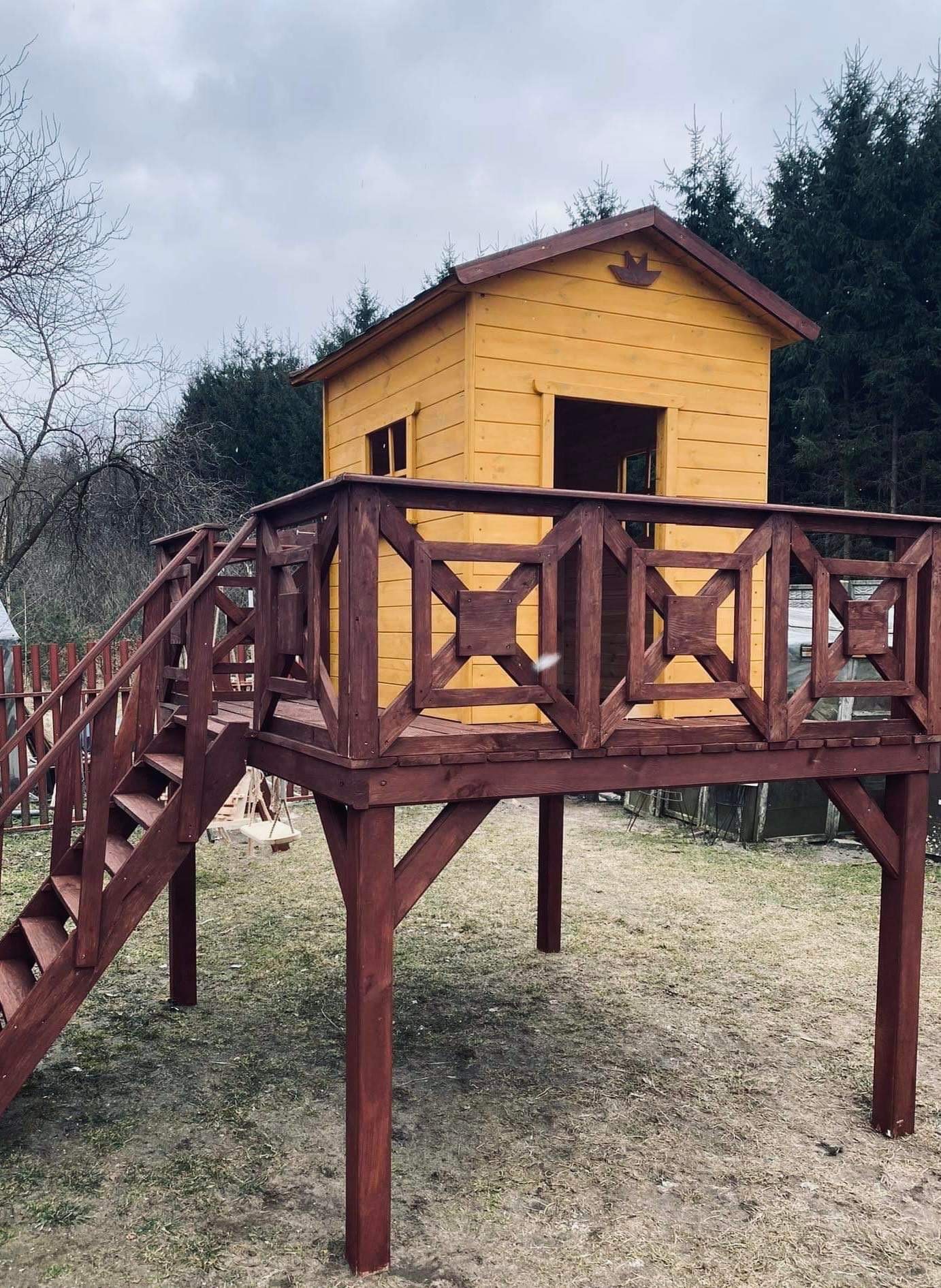 Drewniany plac zabaw w postaci domku na platformie z brązowymi schodami i balustradą, żółty domek z małym ptaszkiem nad wejściem, szary dzień.