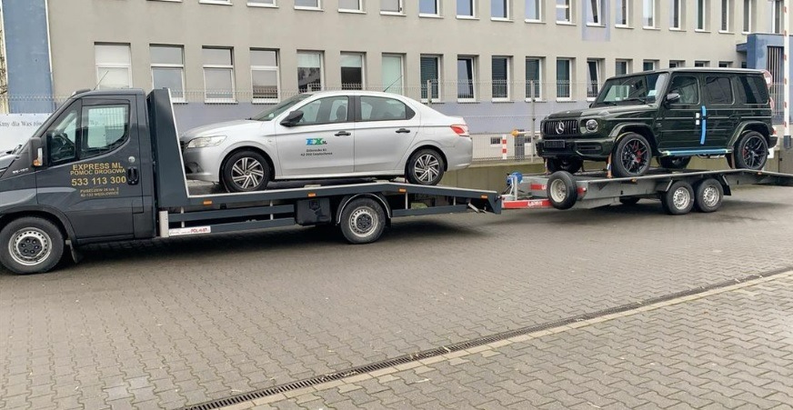 Czarna laweta firmy Express ML przewozi srebrny samochód osobowy i ciemnozielonego Mercedesa G-klasę na przyczepie, na tle budynku z szarymi ścianami i oknami.