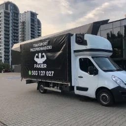 Biała furgonetka z czarną plandeką z napisem 'Transport Przeprowadzki' i logiem bicepsów, zaparkowana na kostce brukowej na tle nowoczesnego budynku biurowego i bloku mieszkalnego.