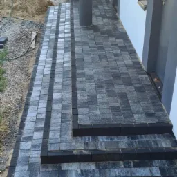 Ścieżka i schody wejściowe do domu wyłożone szarą kostką brukową, z ciemnymi obrzeżami, widoczny fragment trawnika i sterta piasku.