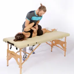 Kobieta leży na beżowym stole do masażu, a mężczyzna w sportowym stroju wykonuje na niej manipulację kręgosłupa lędźwiowego.