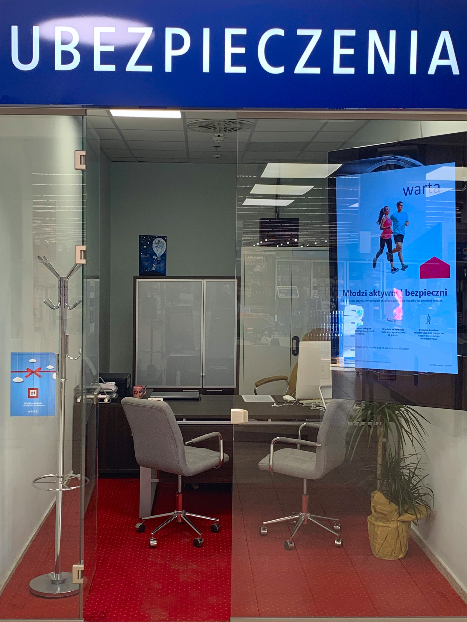 Wnętrze biura ubezpieczeniowego za szklaną witryną z napisem 'UBEZPIECZENIA', widoczne krzesła biurowe, biurko, regał, wieszak na ubrania i plakat reklamowy firmy Warta.