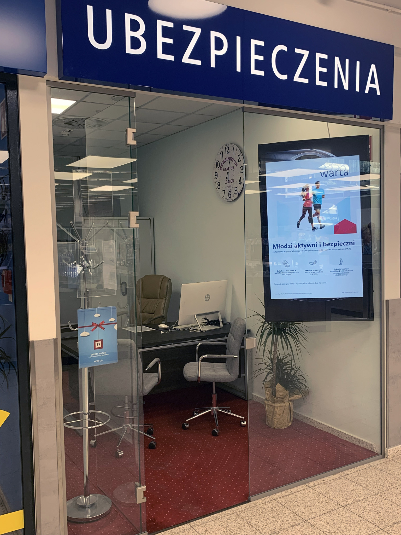 Biuro firmy ubezpieczeniowej Warta z widocznym biurkiem, komputerem, krzesłami i plakatem reklamowym 'Młodzi aktywni i bezpieczni'.