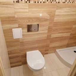 Bedfirm - Nowoczesna toaleta podwieszana z chromowanym panelem spłukującym, otoczona płytkami imitującymi drewno i mozaiką, w narożniku kabina prysznicowa z białym brodzikiem.