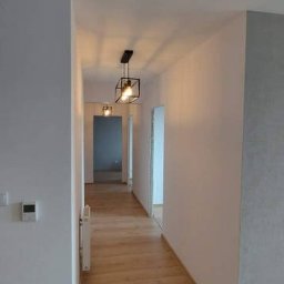 Bedfirm - Długi korytarz w mieszkaniu po remoncie z panelami podłogowymi w odcieniach brązu i beżu, białymi ścianami i minimalistycznymi, czarnymi lampami wiszącymi o geometrycznym kształcie.