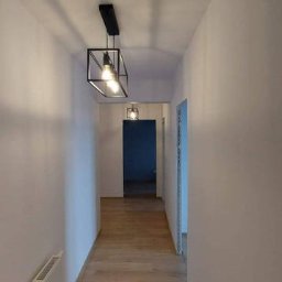 Bedfirm - Długi korytarz z panelami podłogowymi w odcieniach brązu, oświetlony dwoma nowoczesnymi lampami wiszącymi w kształcie czarnych sześcianów z żarówkami Edisona, białe ściany i grzejnik na ścianie po...