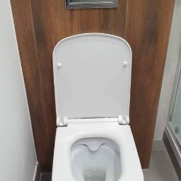 Bedfirm - Biała toaleta wisząca z otwartą klapą na tle drewnianej obudowy ze srebrnym przyciskiem spłukiwania, widoczny fragment szarej podłogi i kabiny prysznicowej.