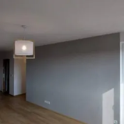 Pusty pokój po remoncie z panelami podłogowymi w odcieniach brązu, szarą ścianą i nowoczesną lampą wiszącą z drewnianą ramą.