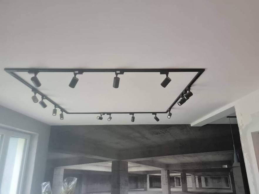 Czarna, geometryczna instalacja oświetleniowa na białym suficie, z reflektorami skierowanymi w różnych kierunkach; ściana z fototapetą przedstawiającą betonową konstrukcję w tle.