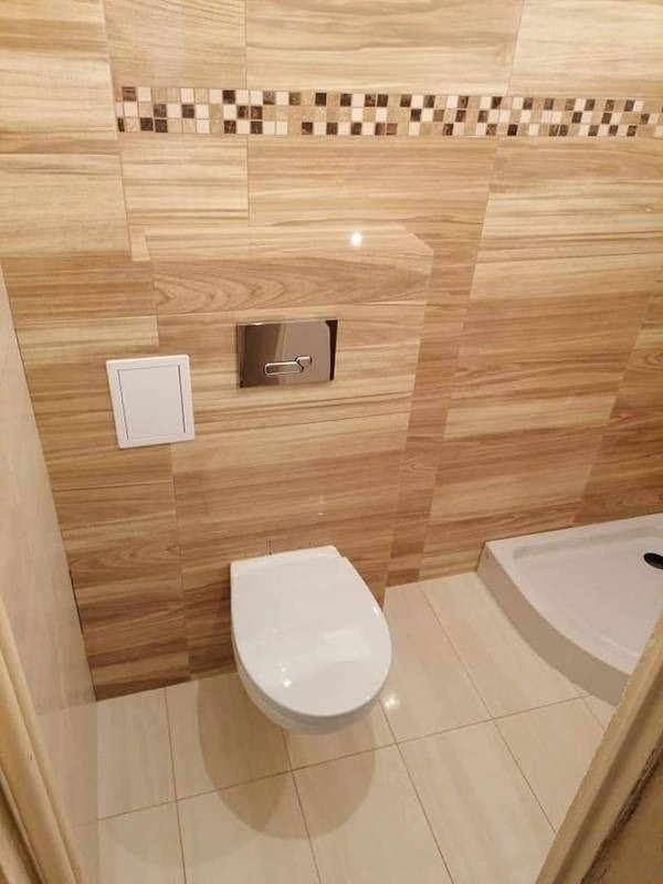 Nowoczesna toaleta podwieszana z chromowanym panelem spłukującym, otoczona płytkami imitującymi drewno i mozaiką, w narożniku kabina prysznicowa z białym brodzikiem.