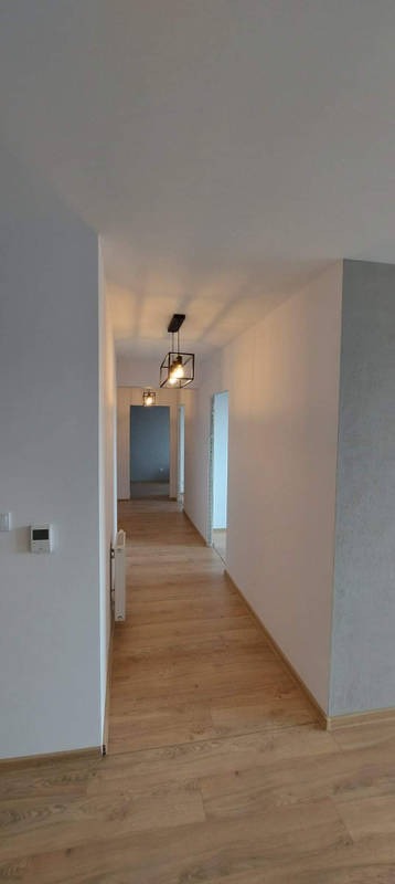 Długi korytarz w mieszkaniu po remoncie z panelami podłogowymi w odcieniach brązu i beżu, białymi ścianami i minimalistycznymi, czarnymi lampami wiszącymi o geometrycznym kształcie.