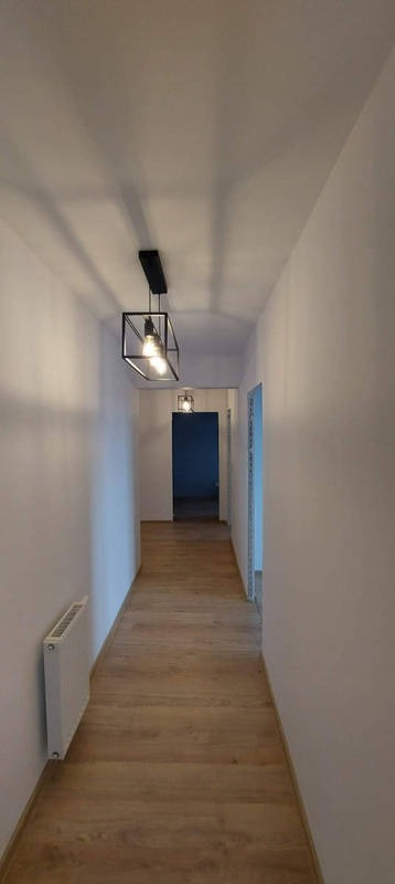 Długi korytarz z panelami podłogowymi w odcieniach brązu, oświetlony dwoma nowoczesnymi lampami wiszącymi w kształcie czarnych sześcianów z żarówkami Edisona, białe ściany i grzejnik na ścianie po...