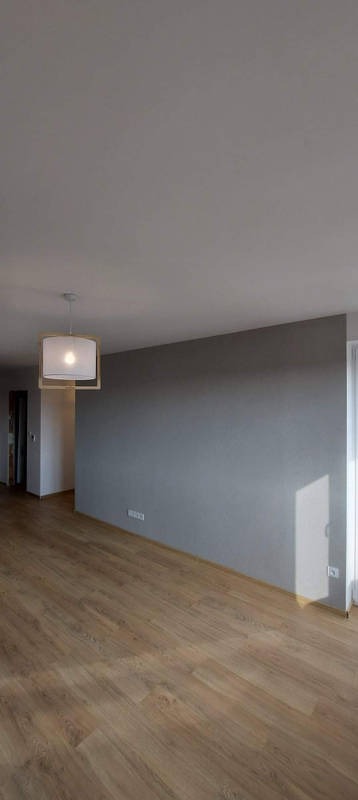 Pusty pokój po remoncie z panelami podłogowymi w odcieniach brązu, szarą ścianą i nowoczesną lampą wiszącą z drewnianą ramą.