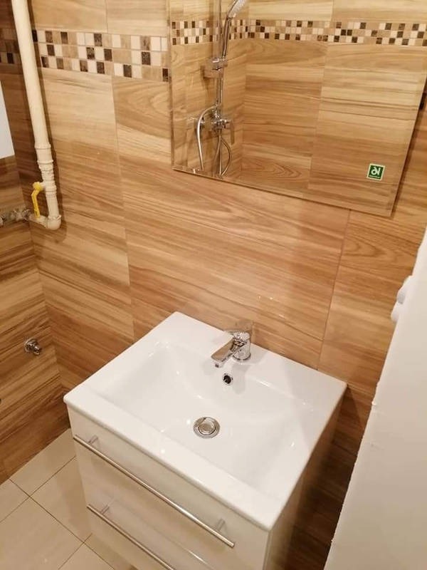 Biała umywalka z szafką i chromowaną baterią na tle ściany wyłożonej płytkami imitującymi drewno, z widoczną rurą gazową po lewej stronie i lustrem z naklejką w prawym górnym rogu.