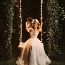 Gajówka Obręb - Para młoda na huśtawce ozdobionej lampkami w ogrodzie, panna młoda w błyszczącej sukni bez ramiączek, pan młody w kamizelce, romantyczna atmosfera o zmierzchu.